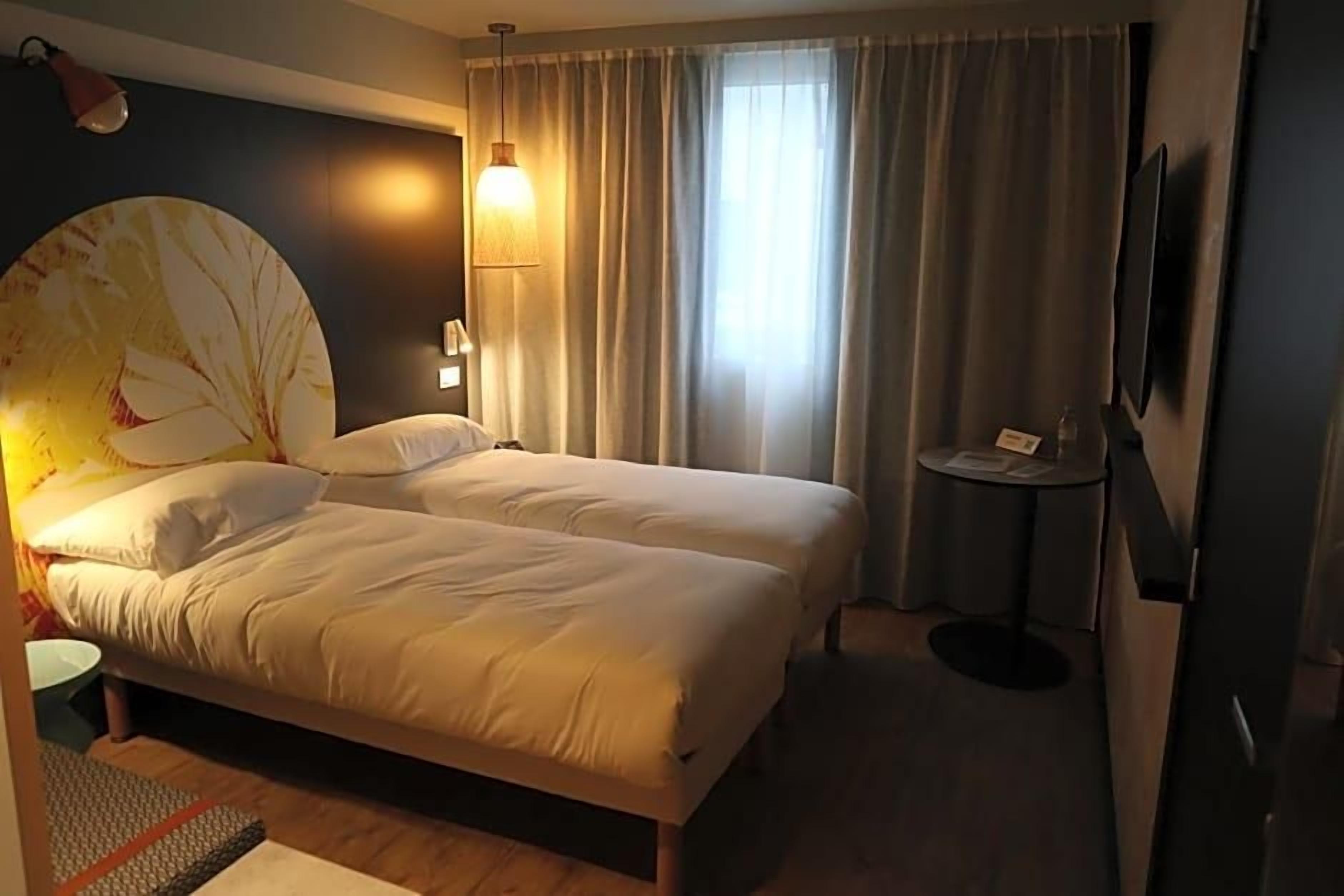 A hotel room with a bed, a small table, a lamp, a TV, and a window with curtains.