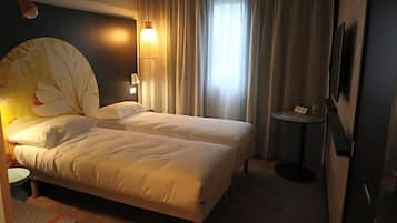 A hotel room with a bed, a small table, a lamp, a TV, and a window with curtains.