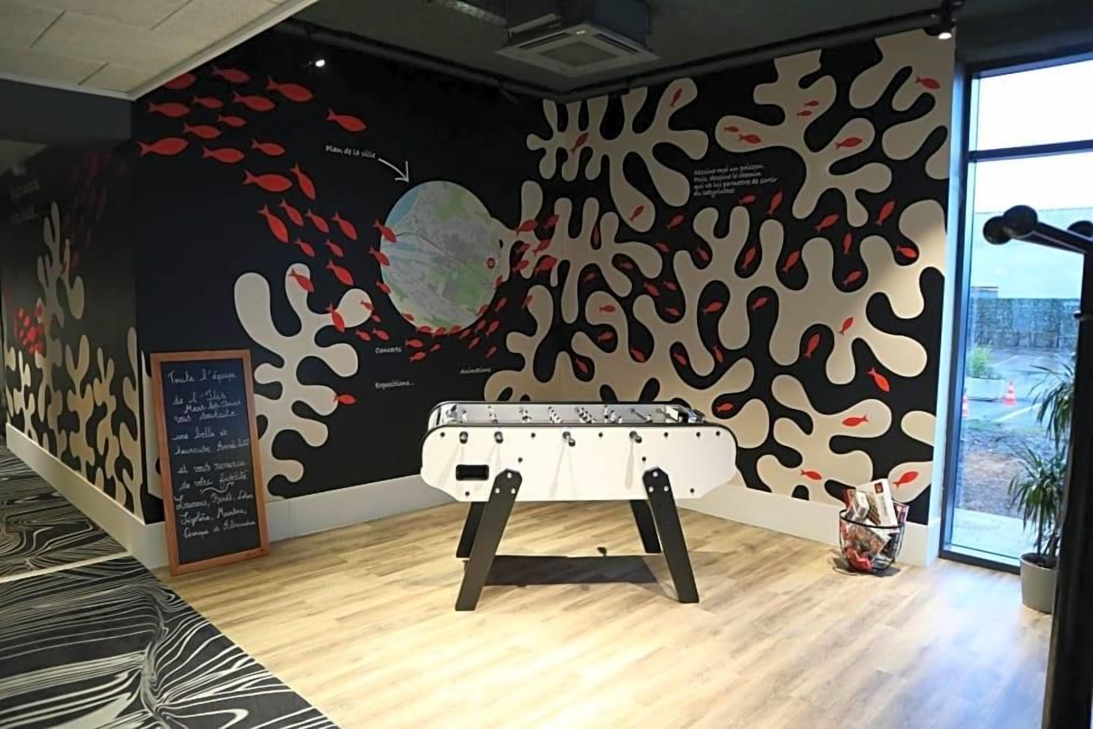 Une pièce avec un baby-foot, une grande fresque murale abstraite et une fenêtre donnant sur un beau paysage.