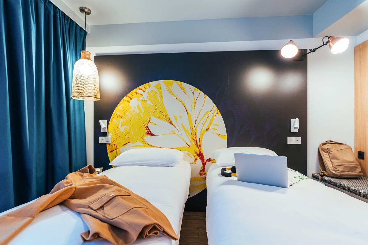A hotel room with two beds, a laptop, a backpack, a wall-mounted lamp, and a large circular artwork.