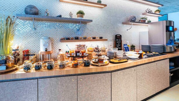 A modern kitchen with a long counter, various food items, and kitchen appliances. - Ibis Styles Le Treport Mers Les Bains (Mers-les-Bains)