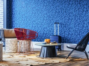 A room with a blue patterned wall, a red chair, a black chair, a suitcase, and a round table with books and a cup. - Ibis Styles Le Treport Mers Les Bains (Mers-les-Bains)