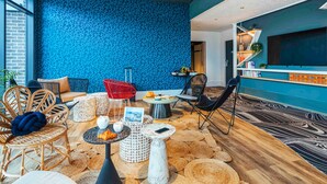 A modern interior with a blue patterned wall, wicker furniture, a wooden floor, and a bar area with books and a TV. - Ibis Styles Le Treport Mers Les Bains (Mers-les-Bains)