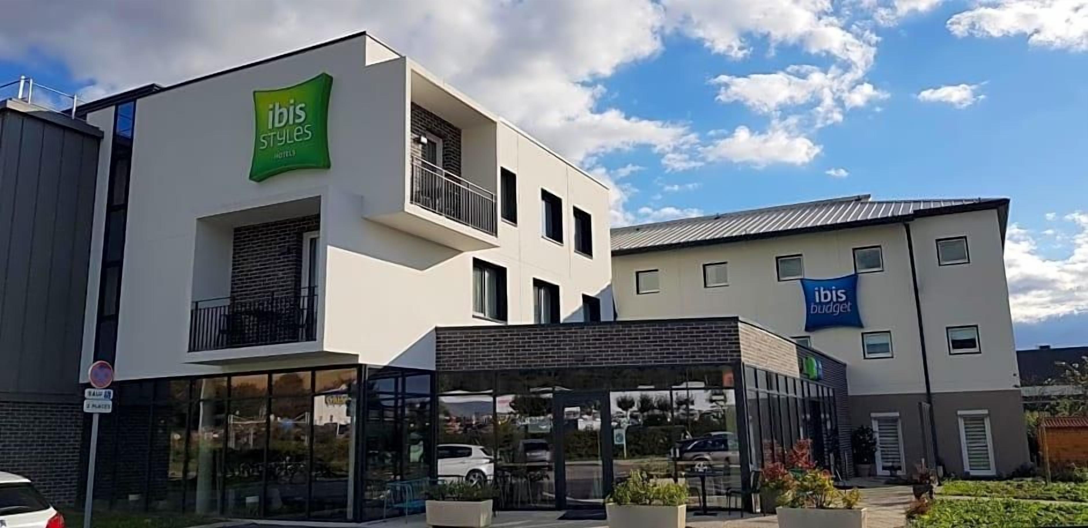 A modern hotel with a green sign, a glass entrance, and a parking area.