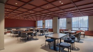 Une salle de conférence spacieuse, dotée de nombreuses tables et chaises, de grandes fenêtres avec des rideaux légers et d’un design de plafond moderne.
