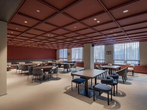 Ein geräumiger Konferenzraum mit mehreren Tischen und Stühlen, großen Fenstern mit leichtem Vorhangstoff und einem modernen Deckenbau.