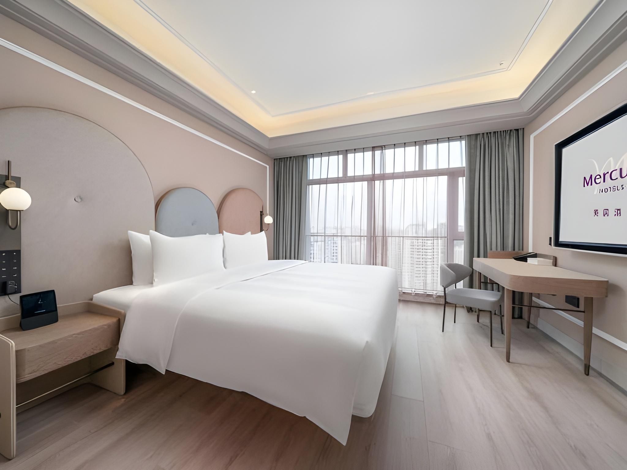 A modern hotel room with a large bed, bedside tables, a desk, and a large window with curtains.