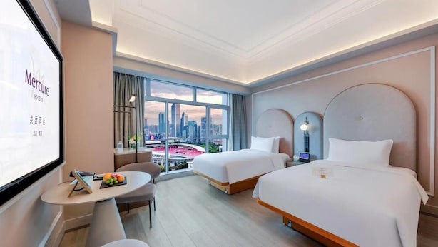 A hotel room with two beds, a desk, a chair, and a large window offering a city view. - Guangzhou Tianhe Sports Center Mercure Hotel (Guangzhou)