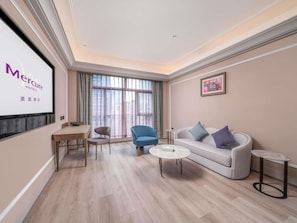 A hotel room with a large window, a sofa, a blue armchair, a small table, and a TV screen displaying hotel branding. - Guangzhou Tianhe Sports Center Mercure Hotel (Guangzhou)