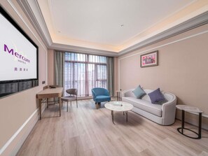 Habitación de hotel con un ventanal grande, un sofá, un sillón azul, una mesita y una pantalla de televisión mostrando la marca del hotel.