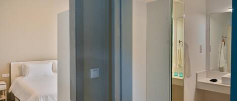 A modern hotel room with a bed, a large mirror, a white bedside table, and a small trash bin.