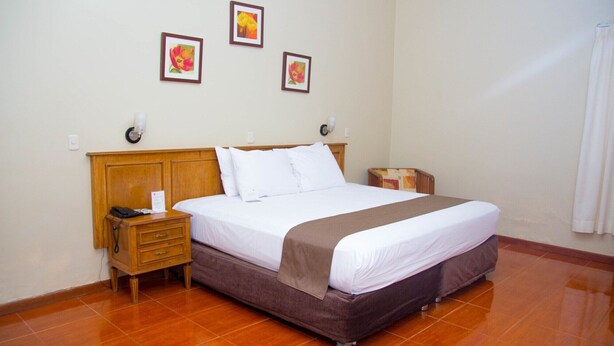 A hotel room with a bed, a nightstand, a lamp, and three framed pictures on the wall. - DM Hoteles Mossone Ica (Ica)
