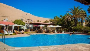 A swimming pool with lounge chairs and umbrellas, surrounded by palm trees and a desert landscape. - DM Hoteles Mossone Ica (Ica)