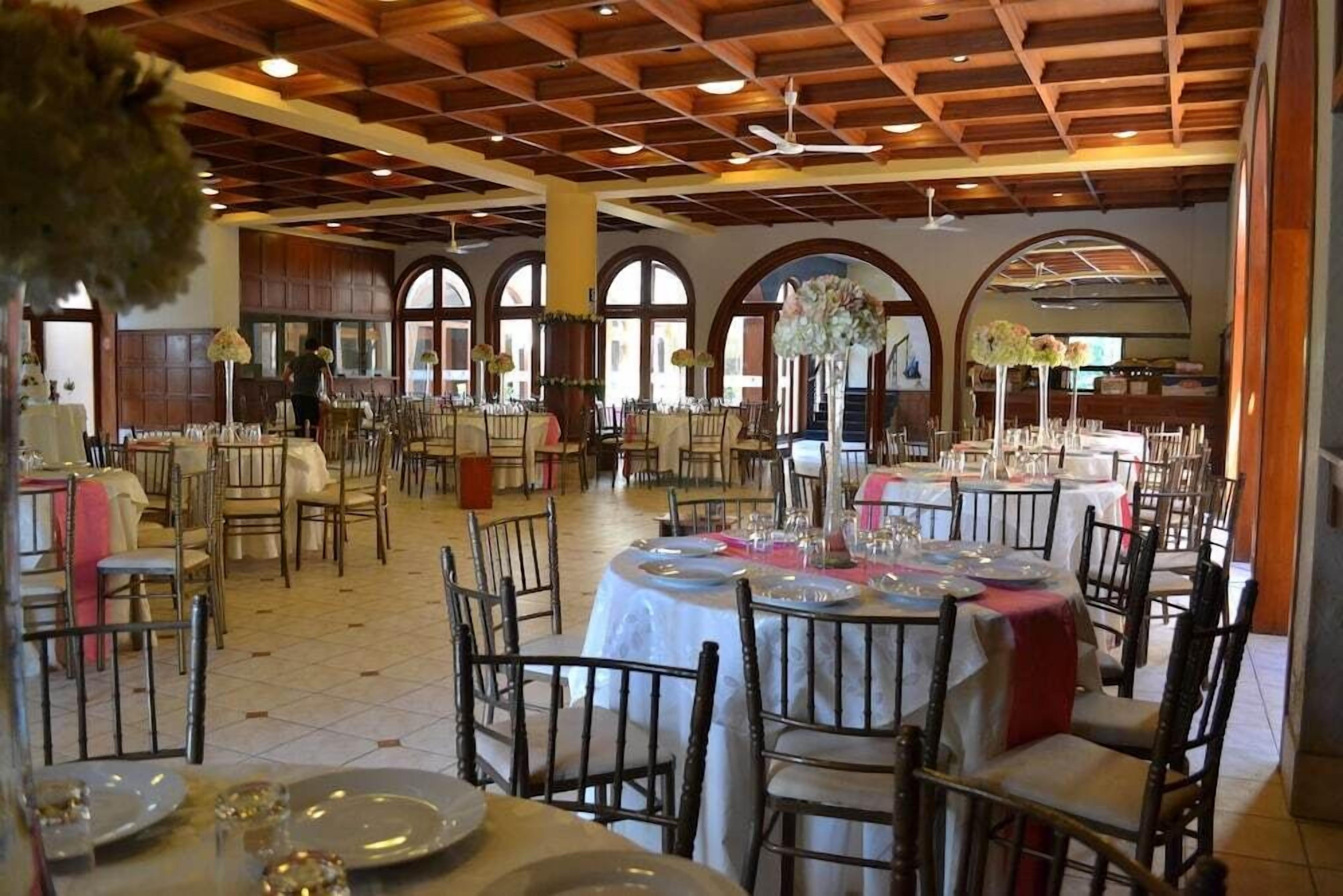 A spacious dining area with multiple round tables, each set with plates, glasses, and cutlery. The room features wooden ceiling beams and large arched windows.