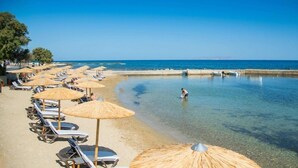 Een strand met ligbedden en rieten parasols, een heldere blauwe zee, en een persoon die in het water waded.