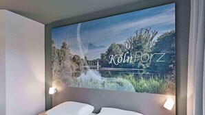 A hotel room with a bed, a wall-mounted artwork of a landscape, and a wall-mounted light fixture. - Ibis Budget Koeln Porz (Cologne)