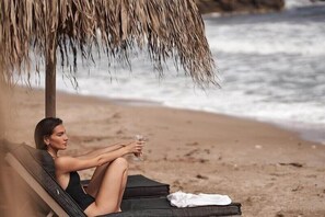 A person reclining on a beach chair under a thatched umbrella, holding a glass. - Nema Design Hotel and Spa - Adults Only (Hersonissos)
