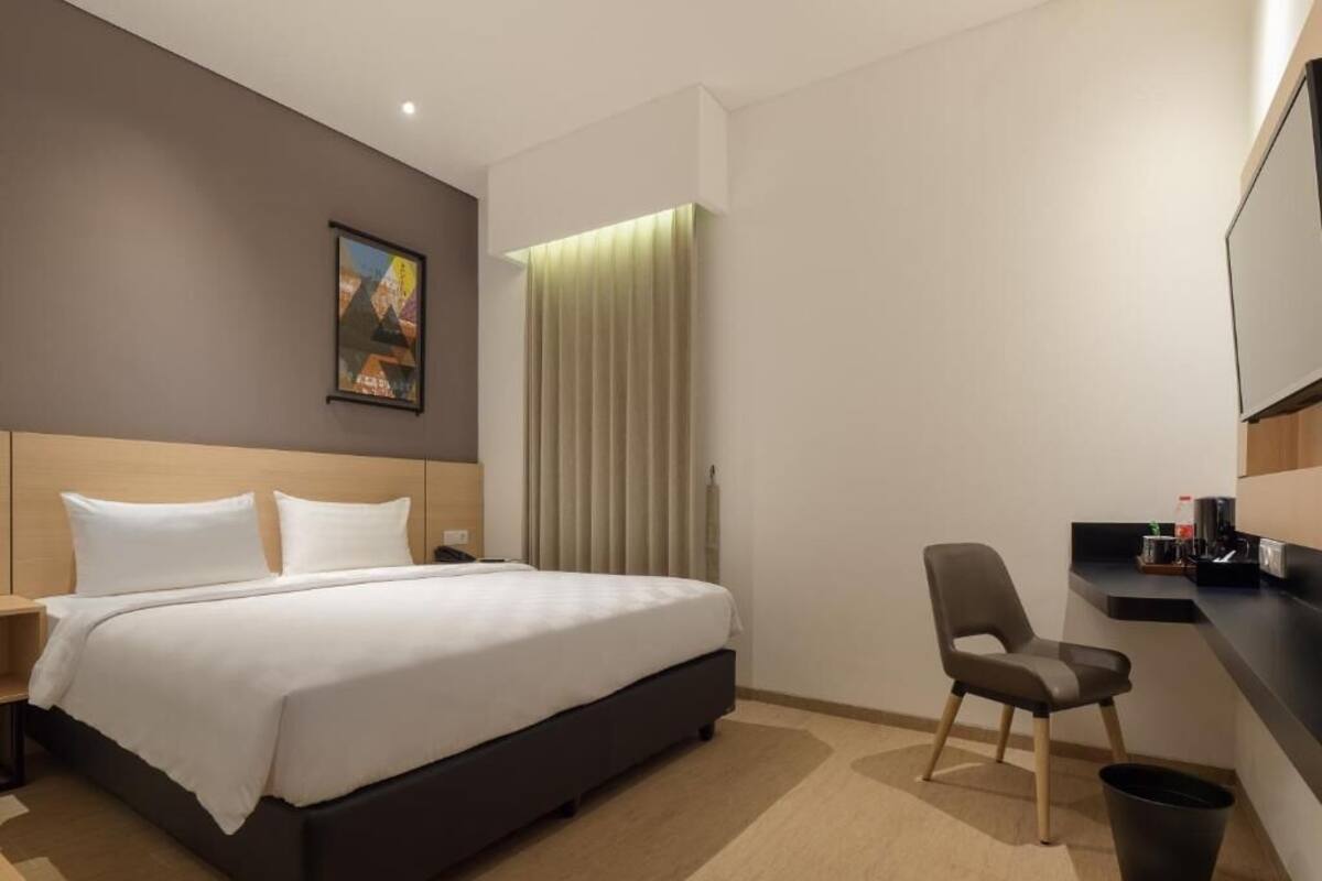 A hotel room with a bed, a chair, a desk, a trash bin, and a painting on the wall.
