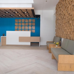 A reception area with a wooden reception desk, a blue wall with wooden panels, a wooden wall with a textured pattern, and a grey sofa with a vase of yellow flowers. - Hotel 88 Blok M (Jakarta)