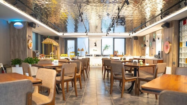 Un restaurante con una decoración moderna, que cuenta con mesas y sillas de madera, una zona de bar central y amplios ventanales que dejan entrar luz natural.