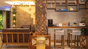Un moderno vestíbulo de hotel con un mostrador de recepción de madera, taburetes de bar, una zona de estar con sofá y una pared decorativa con patrones hexagonales.