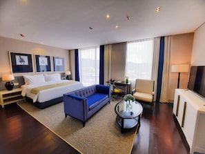 A hotel room with a bed, a blue sofa, a small table with a vase, a chair, a desk with a lamp, and a TV. - Hotel Suni Abepura (Jayapura)