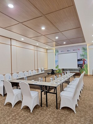 Un salón de conferencias con largas mesas, sillas blancas y una pantalla de proyector.