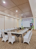 Un salón de conferencias con largas mesas, sillas blancas y una pantalla de proyector.