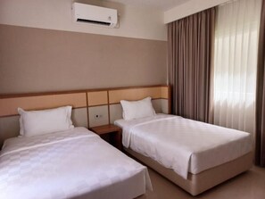 A hotel room with two beds, a headboard, an air conditioning unit, a nightstand, and a window with curtains. - The Allure Villas Managed by Sahid (Pangandaran)