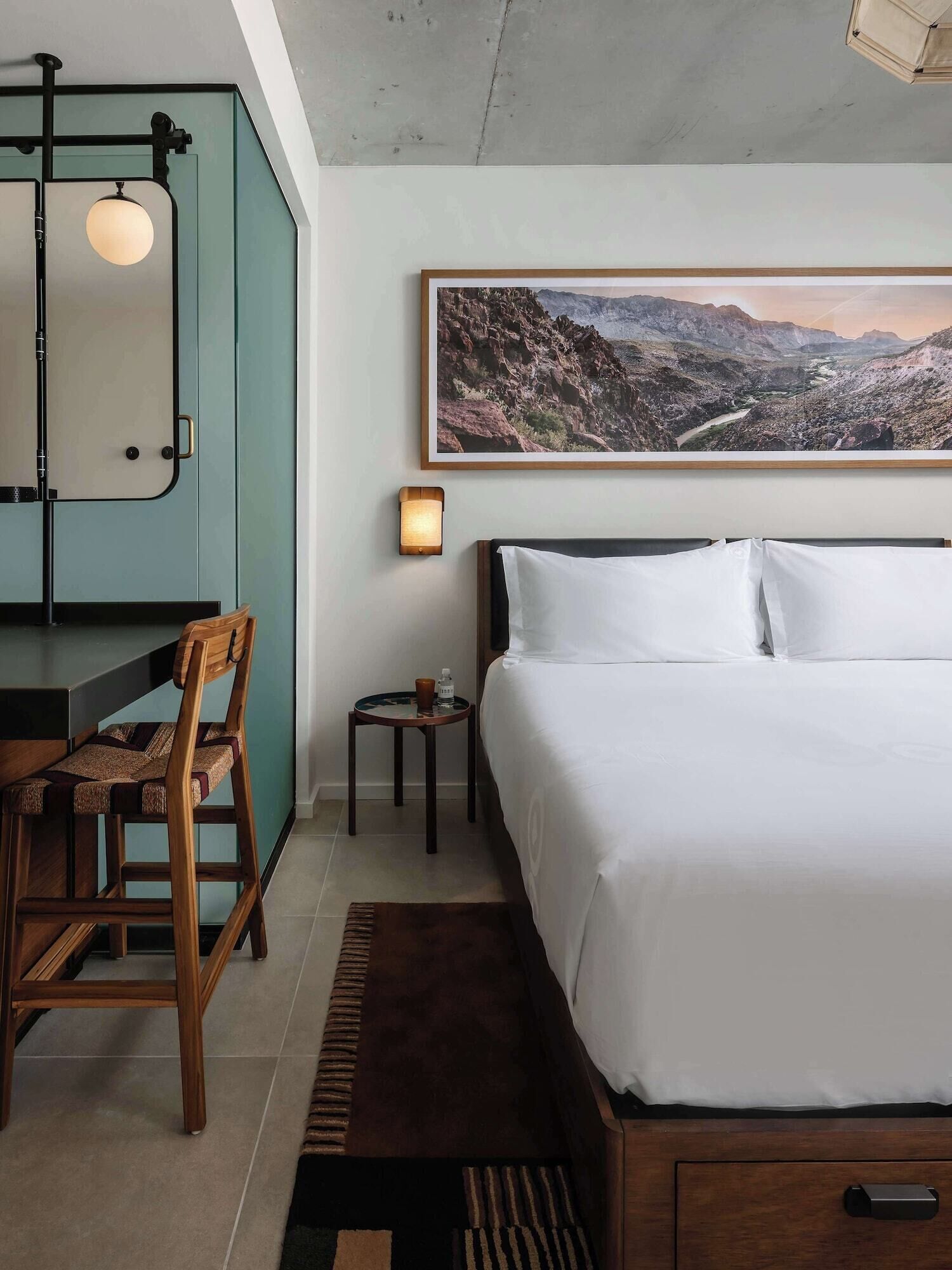 A bedroom with a bed, a desk, a chair, a small table with a lamp, and a framed landscape painting on the wall.