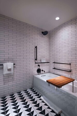 A modern bathroom with a geometric black and white tile floor, a white bathtub with a wooden shelf, and a towel rack with a white towel. - Tommie Austin (Austin)