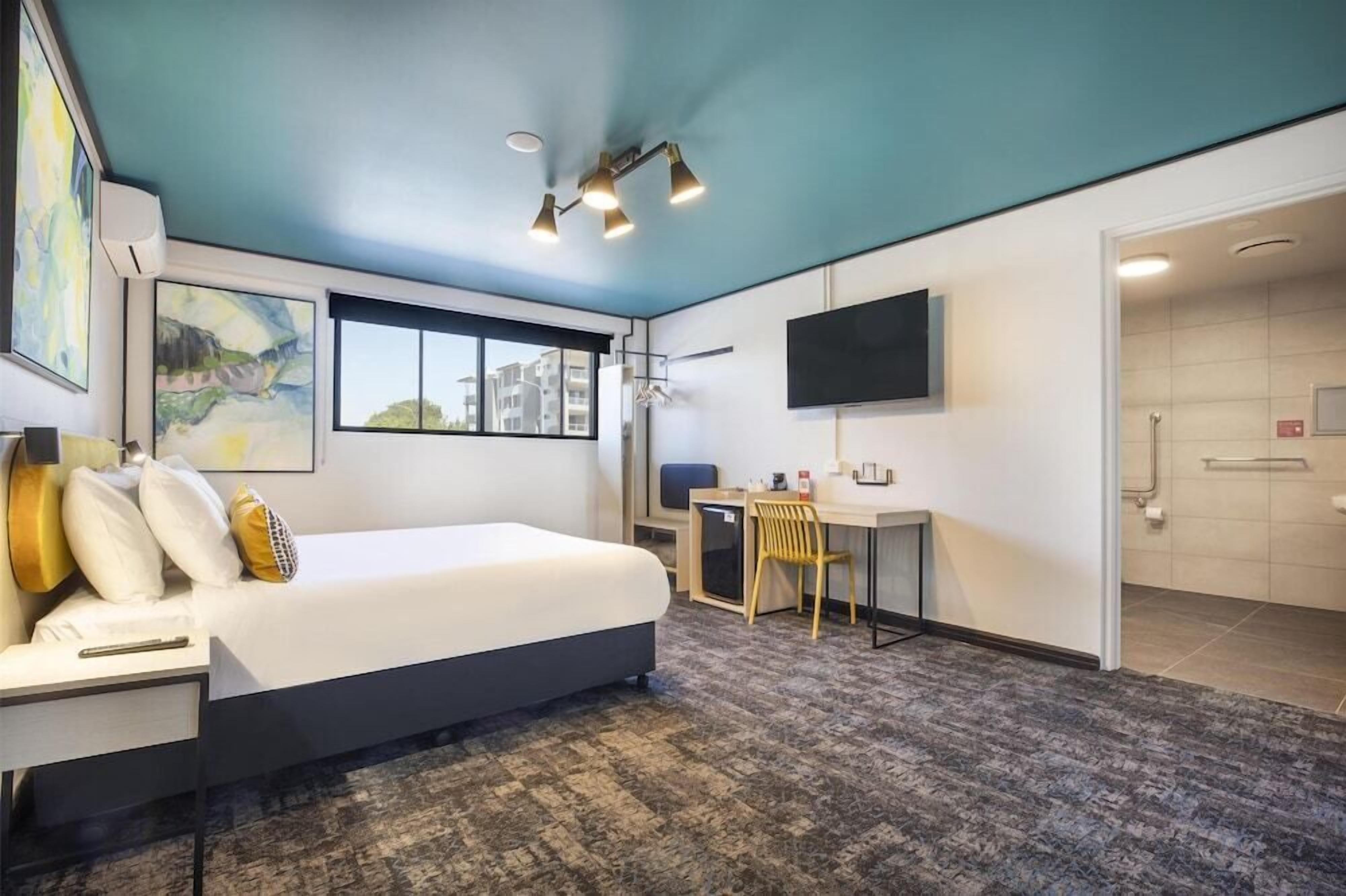 A modern hotel room with a bed, desk, chair, and a TV on the wall.