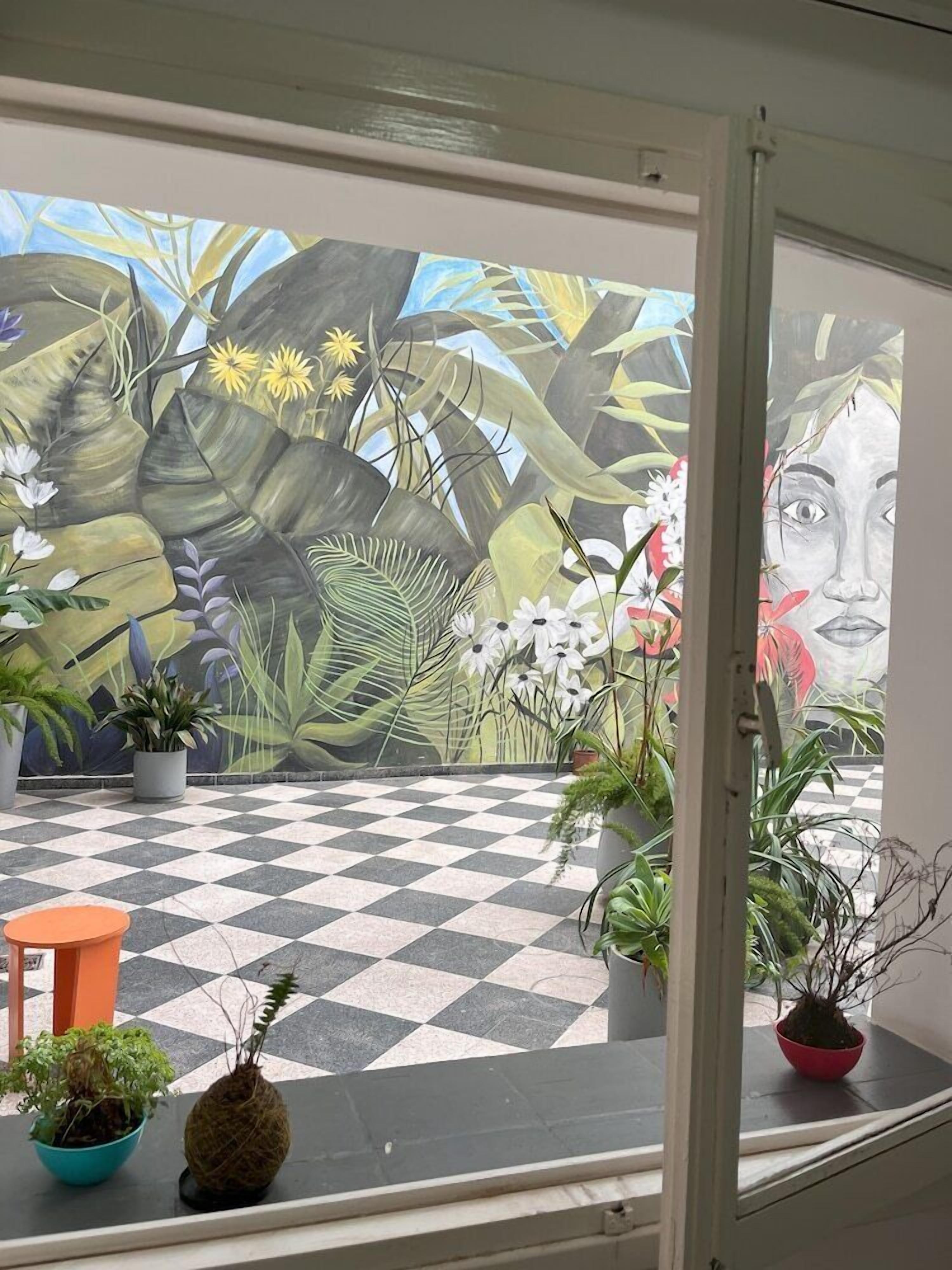 Ein Zimmer mit Schachbrettmuster-Boden, einem großen Fenster mit Blick auf ein Wandgemälde und mehreren Blumentöpfen.