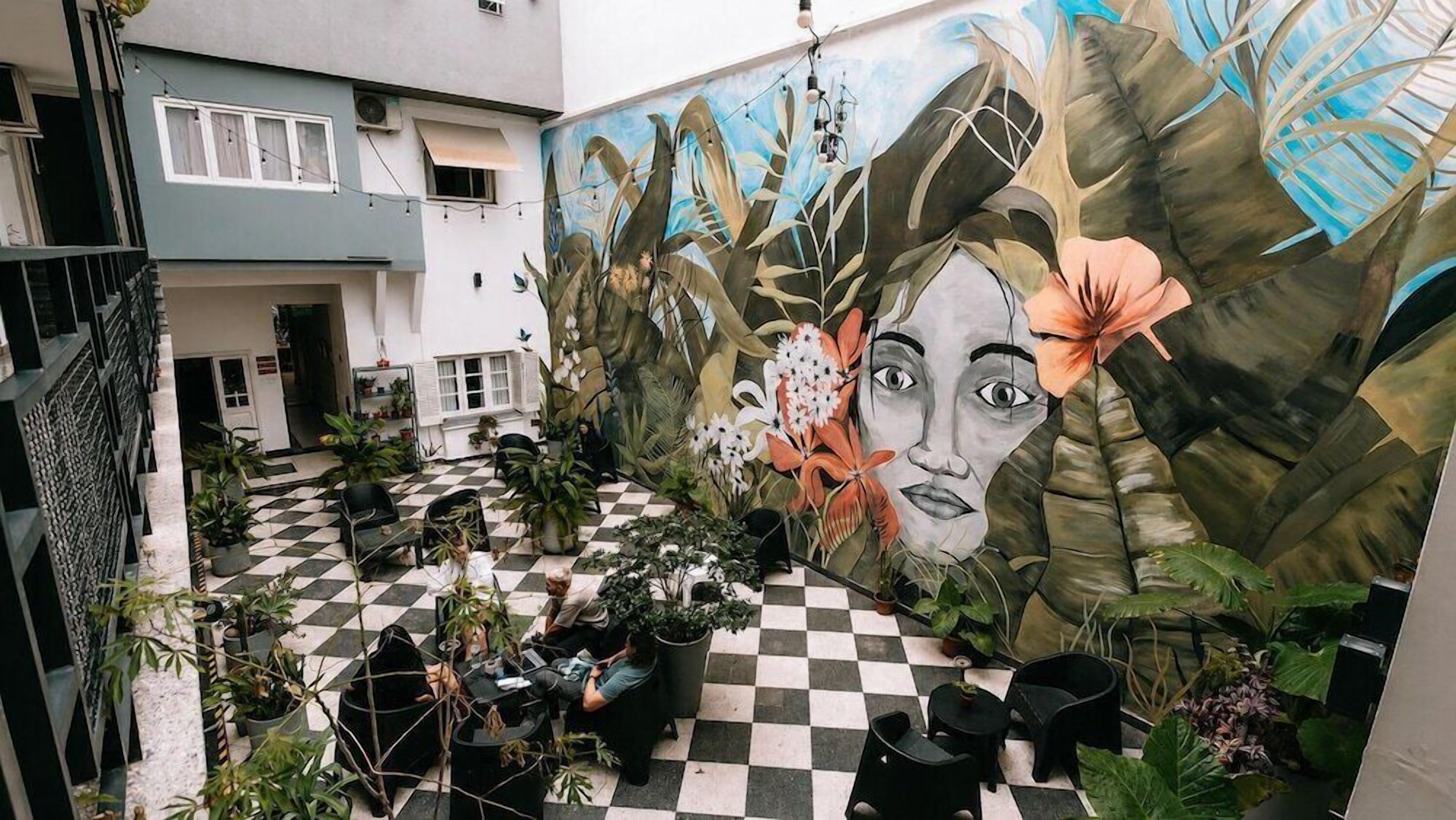 Ein Innenhof mit Schachbrettmuster-Boden, Pflanzkübeln und einer Wandmalerei mit floralen und blattwerkartigen Elementen.