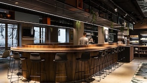 A modern bar area with a curved wooden counter, multiple bar stools, and a high ceiling with exposed beams and lighting. - A-stay Antwerp (Antwerp)