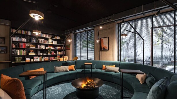 A modern lounge area with a curved green sofa, a central fire pit, a bookshelf, and large windows with blinds. - A-stay Antwerp (Antwerp)