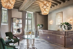 Uma área de recepção com mesa de vidro, duas cadeiras verdes, balcão de recepção de mármore e um candelabro.