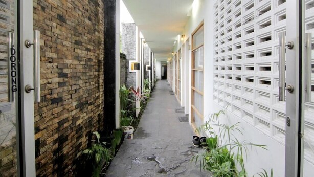 A narrow corridor with a brick wall on one side and a white wall with a grid pattern on the other. There are potted plants along the corridor, and a door labeled "DORONG" is visible. - The Nest Hotel by Zuzu (Depok)