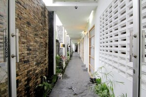 A narrow corridor with a brick wall on one side and a white wall with a grid pattern on the other. There are potted plants along the corridor, and a door labeled "DORONG" is visible. - The Nest Hotel by Zuzu (Depok)
