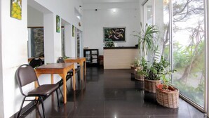 A modern interior with a reception desk, dining area, and large windows offering a view of greenery. - The Nest Hotel by Zuzu (Depok)