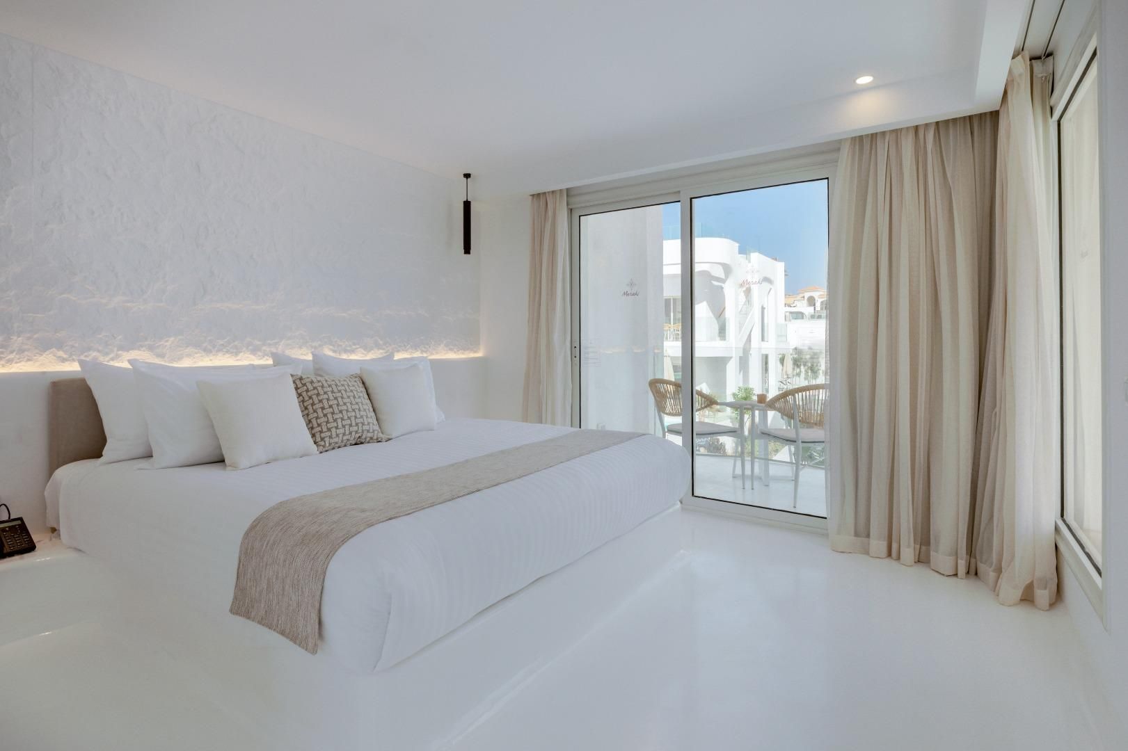 A modern bedroom with a large bed, a sliding glass door to a balcony, and a view of buildings.