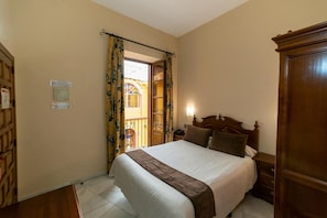A bedroom with a large bed, wooden headboard, and a nightstand. There is a window with curtains, a door leading to an outdoor area, and a wooden wardrobe. - Hotel Mezquita (Córdoba)