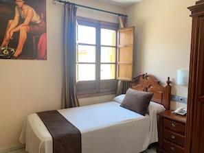 A hotel room with a bed, a wooden nightstand with a phone, a window with curtains, and a painting on the wall. - Hotel Mezquita (Córdoba)