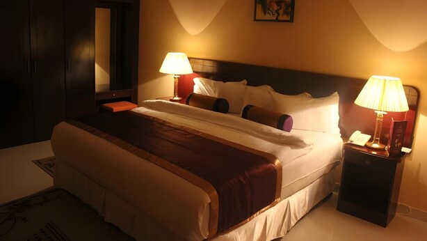 A hotel room with a large bed, two bedside lamps, a nightstand with a phone, and a painting on the wall. - Royal Garden Hotel (Sohar)