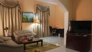 A living room with a sofa, coffee table, and a TV. There is a desk with a chair and a lamp. The room has curtains and a painting on the wall. - Royal Garden Hotel (Sohar)