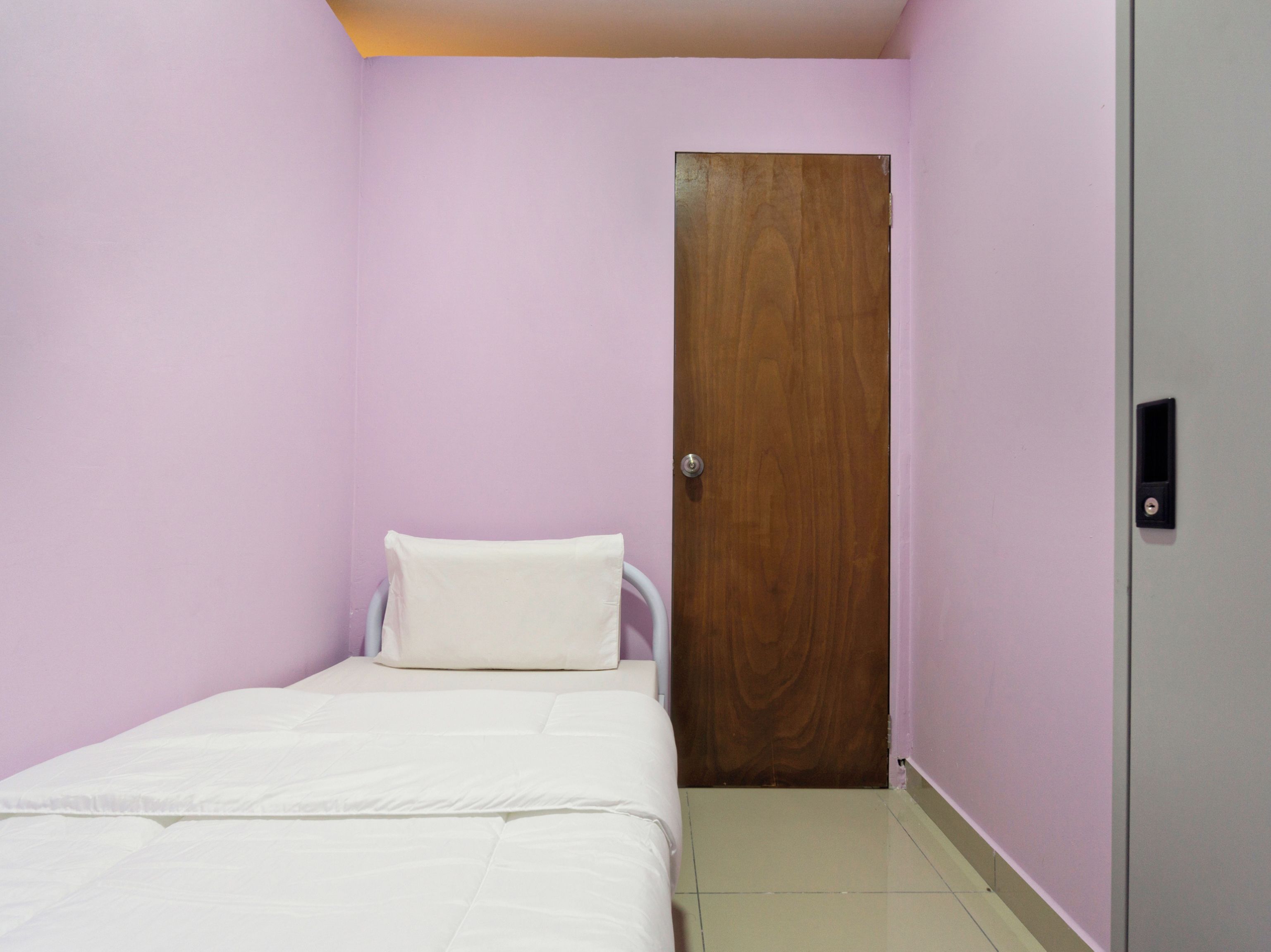 Pequeña habitación con una cama individual, puerta de madera y cubrecama blanco.