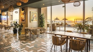 A restaurant with a herringbone floor pattern, wicker chairs, and tables. It features large windows with a view of a sunset and a distant cityscape. - Bavaro Beach Dakhla (Dakhla)