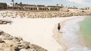 A beach with a person walking along the shore, a rocky breakwater, and a row of buildings in the background. - Bavaro Beach Dakhla (Dakhla)