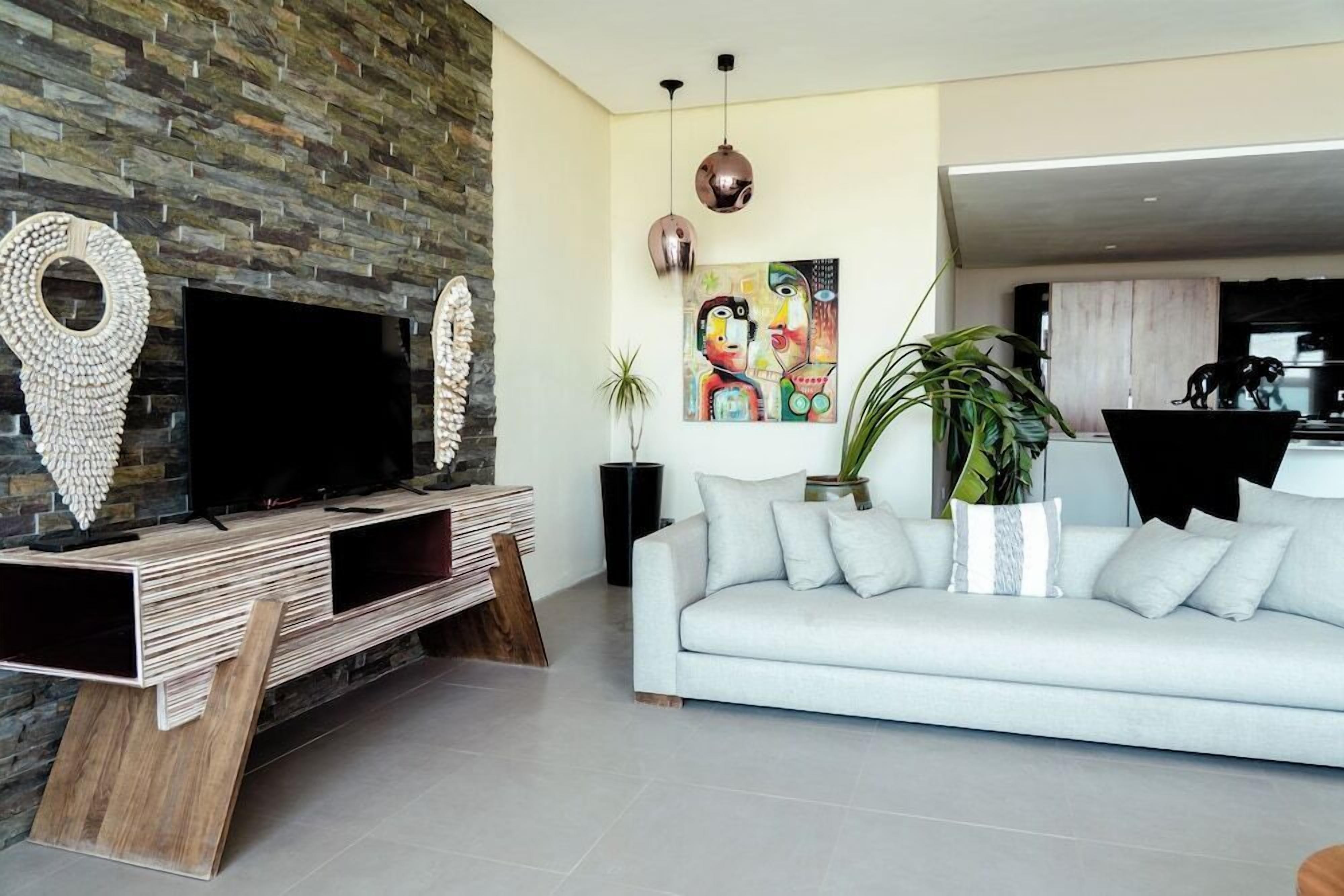 A modern living room with a stone wall, a flat-screen TV, a light grey sofa, and a colorful abstract painting.