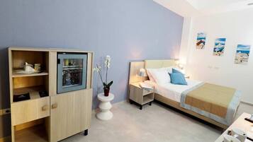 A hotel room with a bed, a bedside table with a lamp, a small white table with a vase of flowers, a wardrobe, and a shelf with various items.